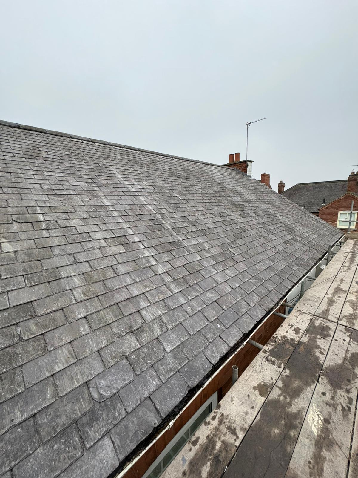 Grey slate roof seen along the pitch from scaffolding, with chimneys and terraced houses behind
