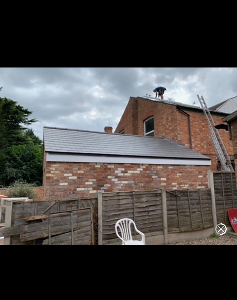 Newly tiled lean-to roof on a brick outbuilding with a roofer working on the house roof behind