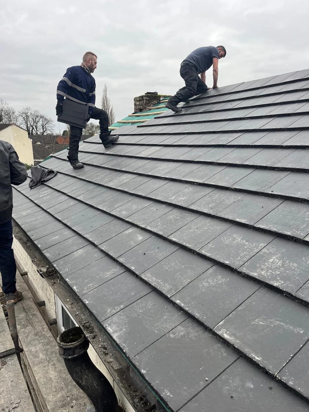 Two roofers fitting dark tiles across a roof slope