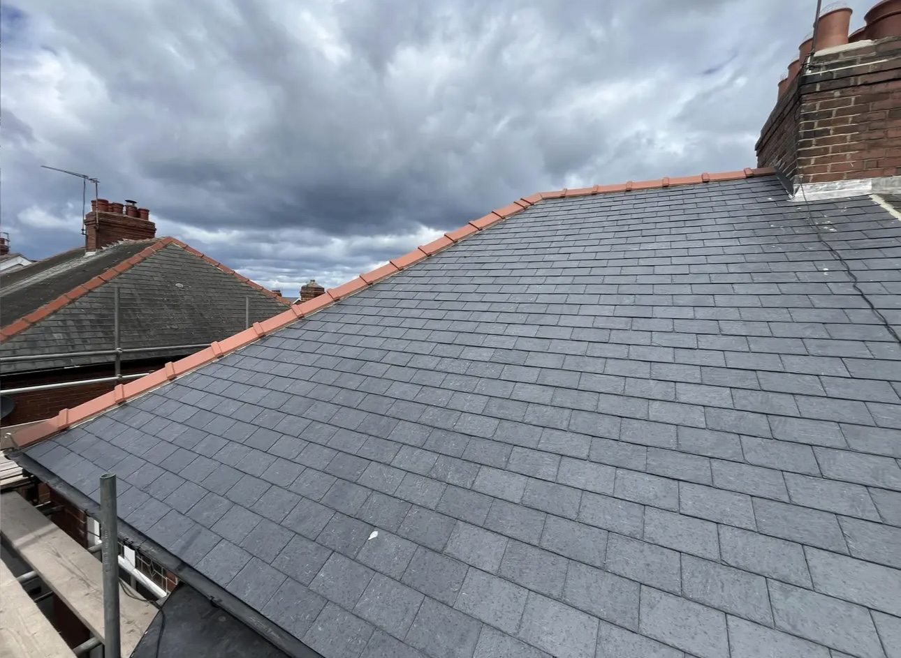 Grey slate roof topped with red ridge tiles, chimneys and neighbouring rooftops behind